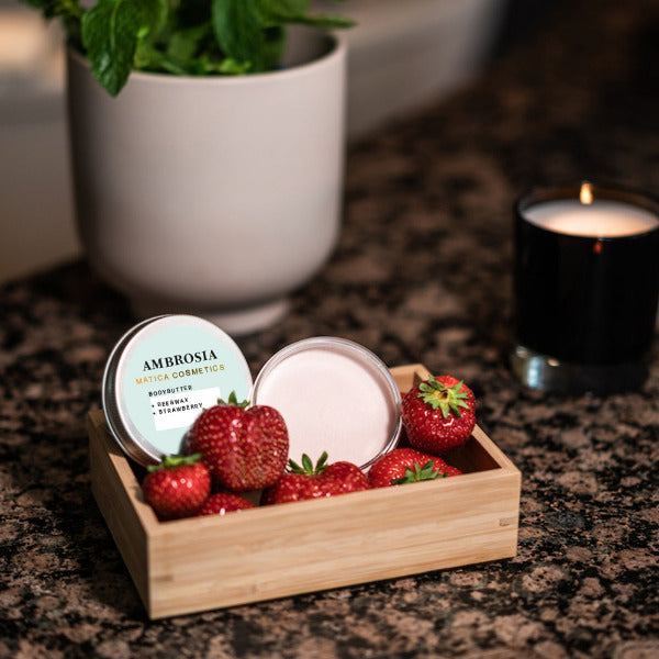 Bodybutter Ambrosia - Erdbeere von Matica Cosmetics in einer Holzschale, umgeben von frischen Erdbeeren und einer flackernden Kerze – für ein sinnliches Pflegeerlebnis.